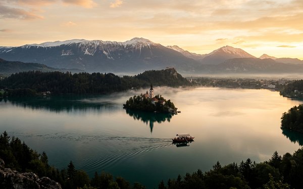 Lake Bled at sunrise with its island church, a small boat crossing calm water, and misty Julian Alps behind.